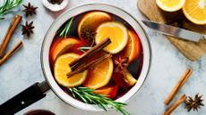 overhead shot of a saucepan filled with red wine cinnamon sticks, rosemary sprigs and lots of sliced orange